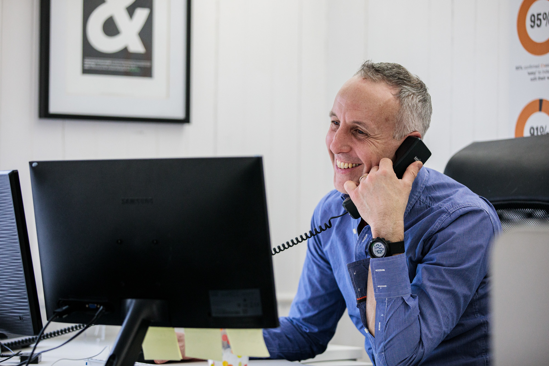 A man sat at a desk, on the phone smiling.